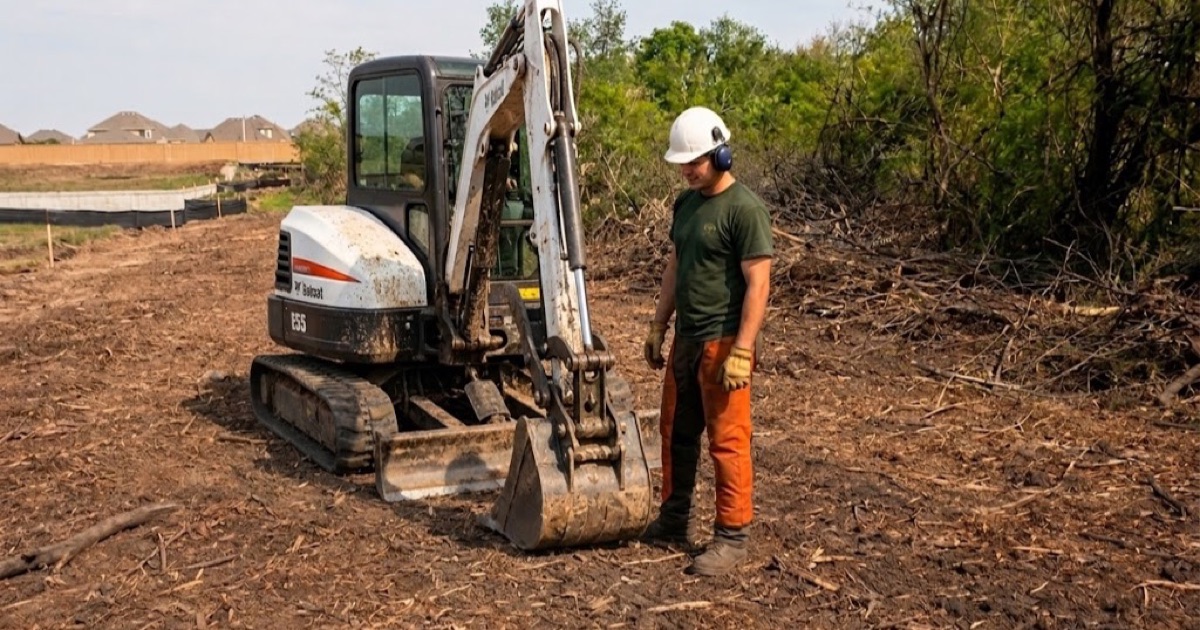 Worker swinging the parked grapple arm of a mulching machine onto a honey locust cull log at the windrow on a Spring Hill site-prep job.